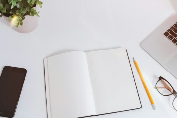 An open empty notebook on a white desk next to an iPhone and a MacBook