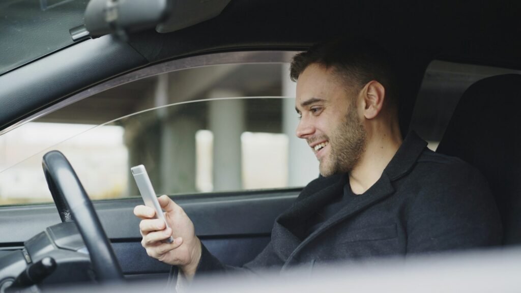 A smiling man sitting in a parked car and looking at his mobile phone