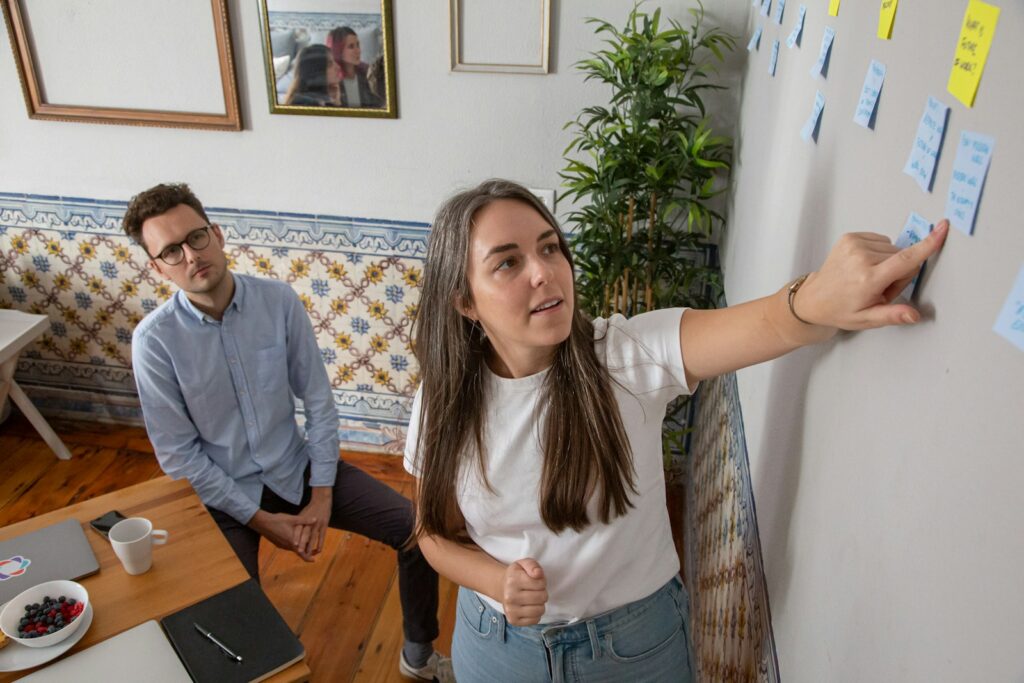 A woman points at sticky notes on a wall while a seated colleague listens