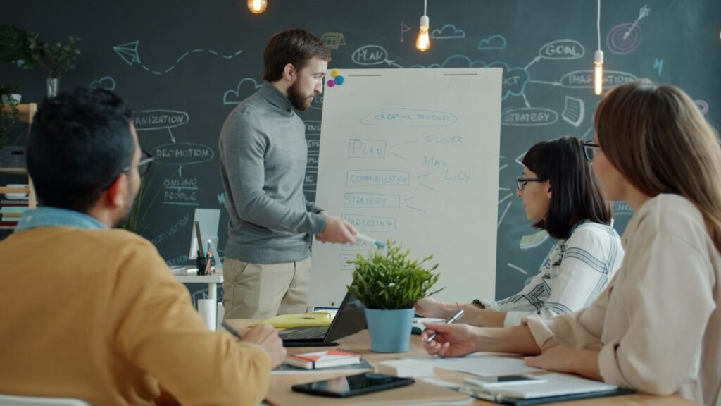 A man presenting a plan on a whiteboard to colleagues seated around an office table