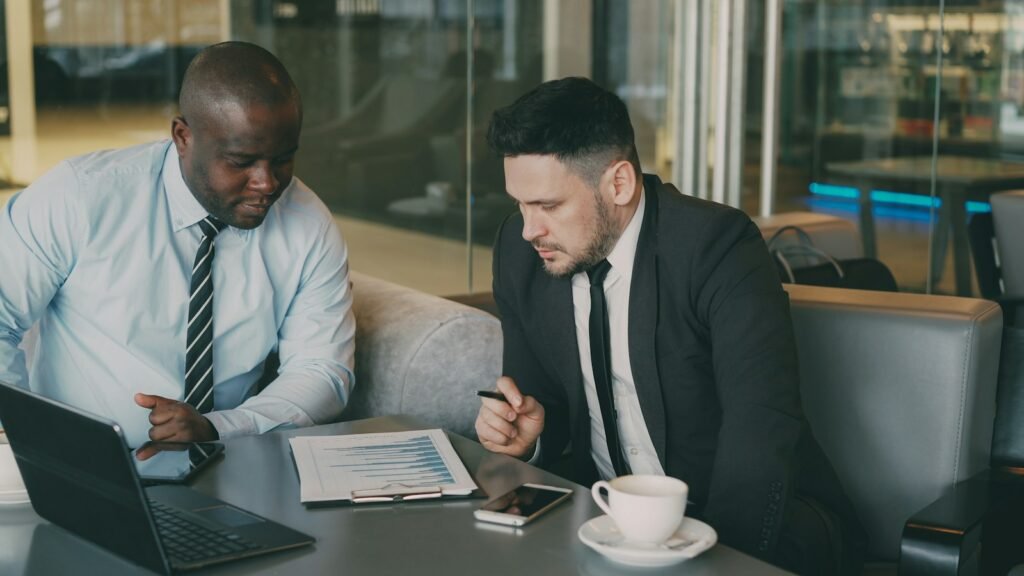 Two businessmen reviewing printed charts beside a laptop and coffee at a cafe table