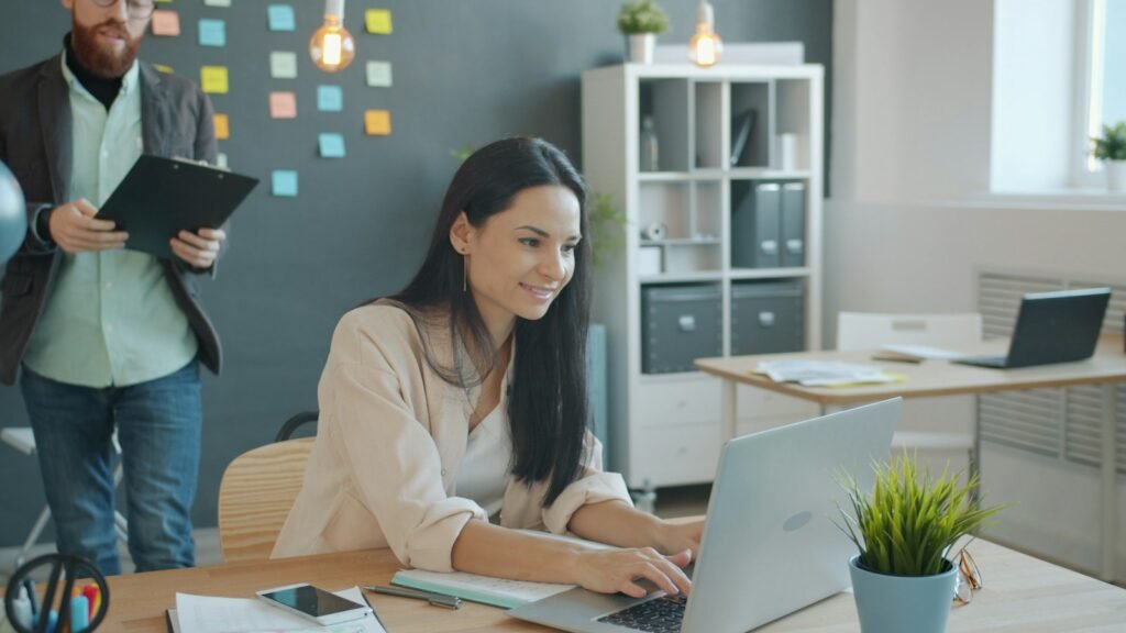 A smiling woman working on a laptop while a colleague holding a clipboard walks past